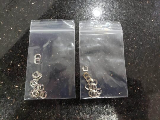 Two clear plastic bags on a dark surface, each holding several small, silver, rectangular Internal Click Plates.